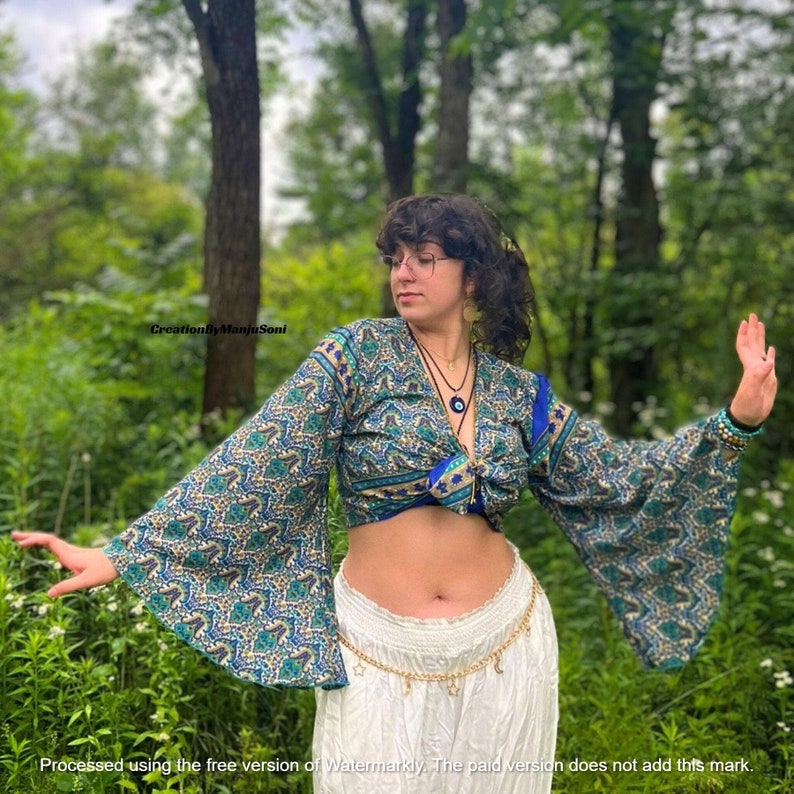 May include: A woman wearing a blue and green patterned crop top with bell sleeves, a white skirt, and a gold belt with charms. She is standing in a forest setting.