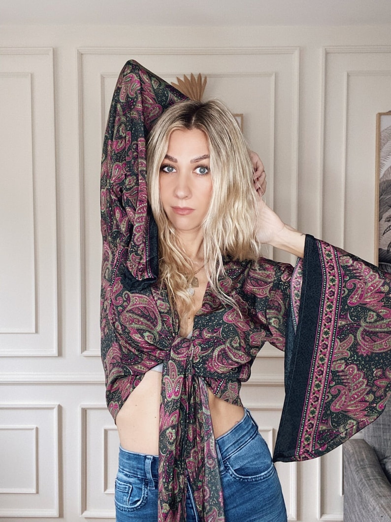 May include: A woman wearing a black and pink paisley print crop top with long sleeves and a tie front. The top has a deep V-neckline and is tied at the waist. The woman is wearing blue jeans.