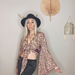 May include: A woman wearing a tie-front top with a paisley print and bell sleeves. The top is tied at the waist and has a burgundy and cream color scheme. She is also wearing a black hat and dark denim jeans.