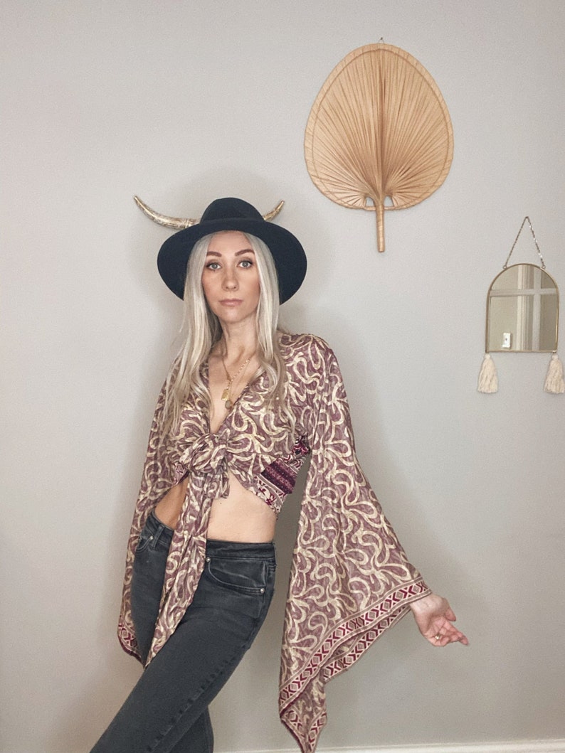 May include: A woman wearing a tie-front top with a paisley print and bell sleeves. The top is tied at the waist and has a burgundy and cream color scheme. She is also wearing a black hat and dark denim jeans.