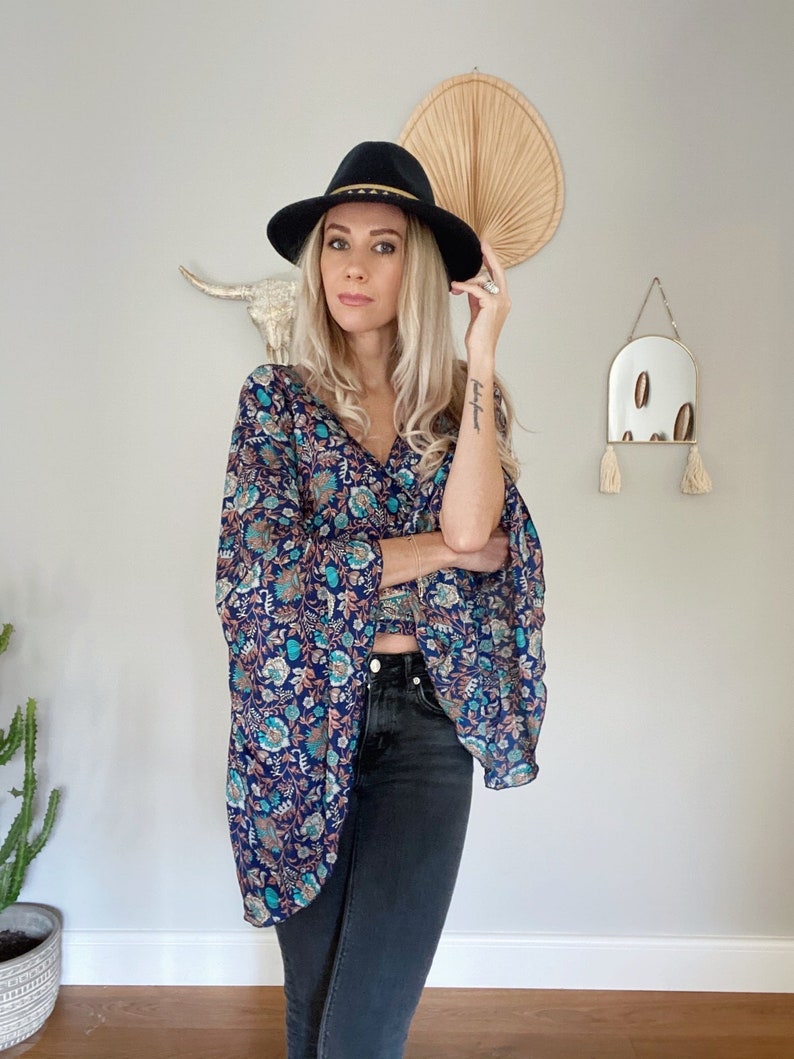 May include: A woman wearing a blue and floral print top with a black hat. The top has a loose fit and long sleeves. She is also wearing black jeans.