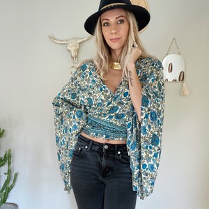 May include: A woman wearing a blue and white floral print wrap top with bell sleeves, black jeans, and a black fedora with a gold band.