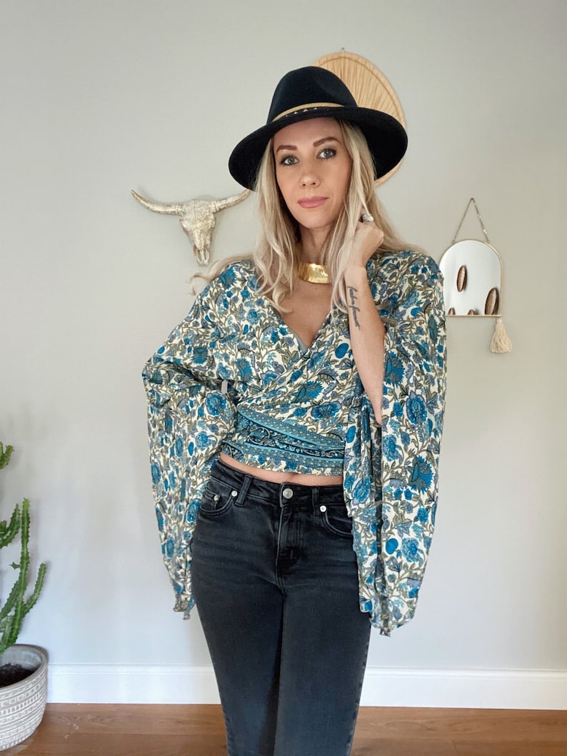 May include: A woman wearing a blue and white floral print wrap top with bell sleeves, black jeans, and a black fedora with a gold band.