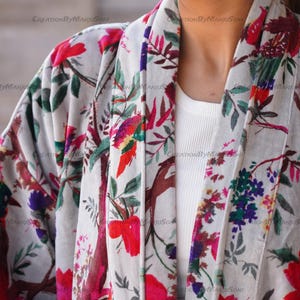 May include: A white kimono with a floral print featuring pink, red, green, and purple flowers. The kimono has a long sleeve and a wide collar.