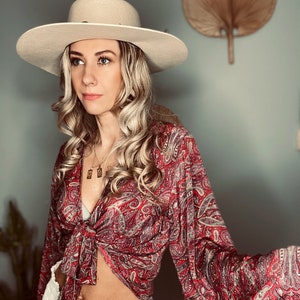 May include: A woman wearing a red and white paisley print blouse tied at the waist, white pants, and a wide-brimmed beige hat.