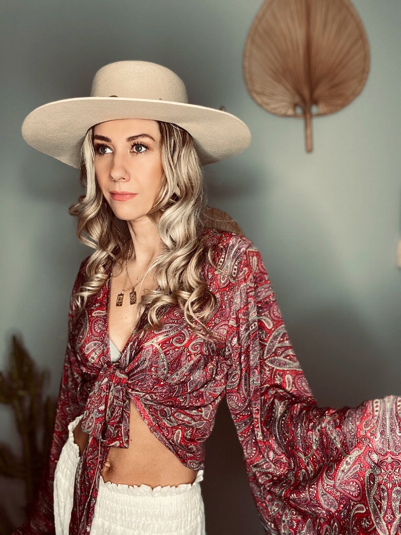May include: A woman wearing a red and white paisley print blouse tied at the waist, white pants, and a wide-brimmed beige hat.