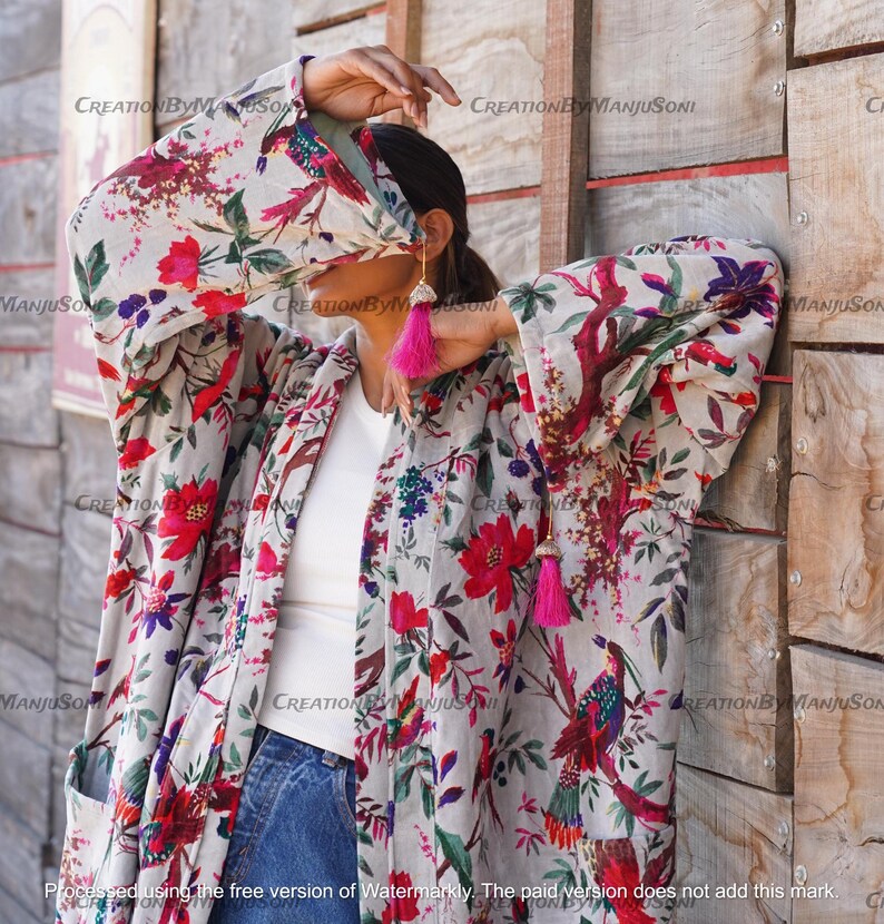 May include: A long-sleeved, white kimono with a floral and bird print. The kimono has a pink tassel on the left side and a pink tassel on the right side. The kimono is worn over a white t-shirt and blue jeans.