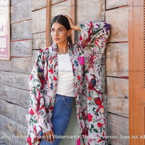 May include: A white kimono with a floral print featuring birds and flowers. The kimono has a long, flowing silhouette and is made of a lightweight fabric. The model is wearing the kimono over a white t-shirt and blue jeans.