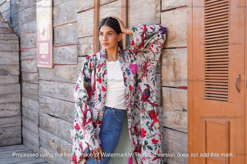May include: A white kimono with a floral print featuring birds and flowers. The kimono has a long, flowing silhouette and is made of a lightweight fabric. The model is wearing the kimono over a white t-shirt and blue jeans.