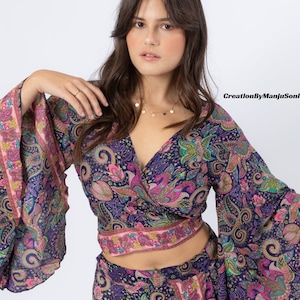 May include: A woman wearing a blue and pink paisley print wrap top with long bell sleeves and a matching skirt. The top is cropped and the skirt is long. She is wearing a gold necklace with small circular pendants.