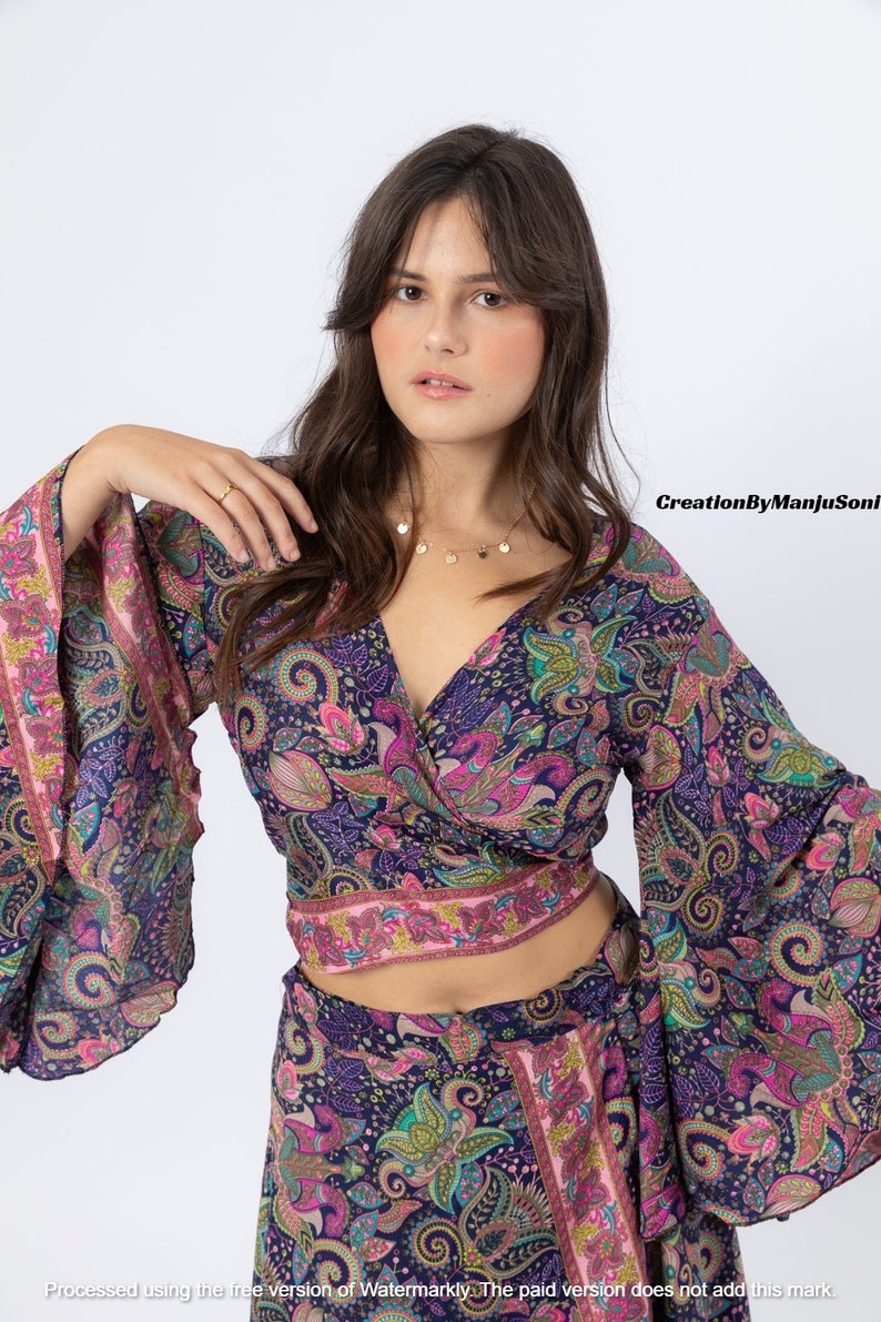 May include: A woman wearing a blue and pink paisley print wrap top with long bell sleeves and a matching skirt. The top is cropped and the skirt is long. She is wearing a gold necklace with small circular pendants.