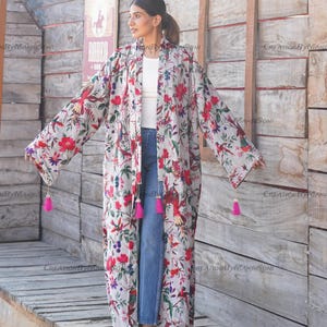 May include: A long, white floral print kimono with pink tassels on the sleeves. The kimono is worn over a white t-shirt and blue jeans.