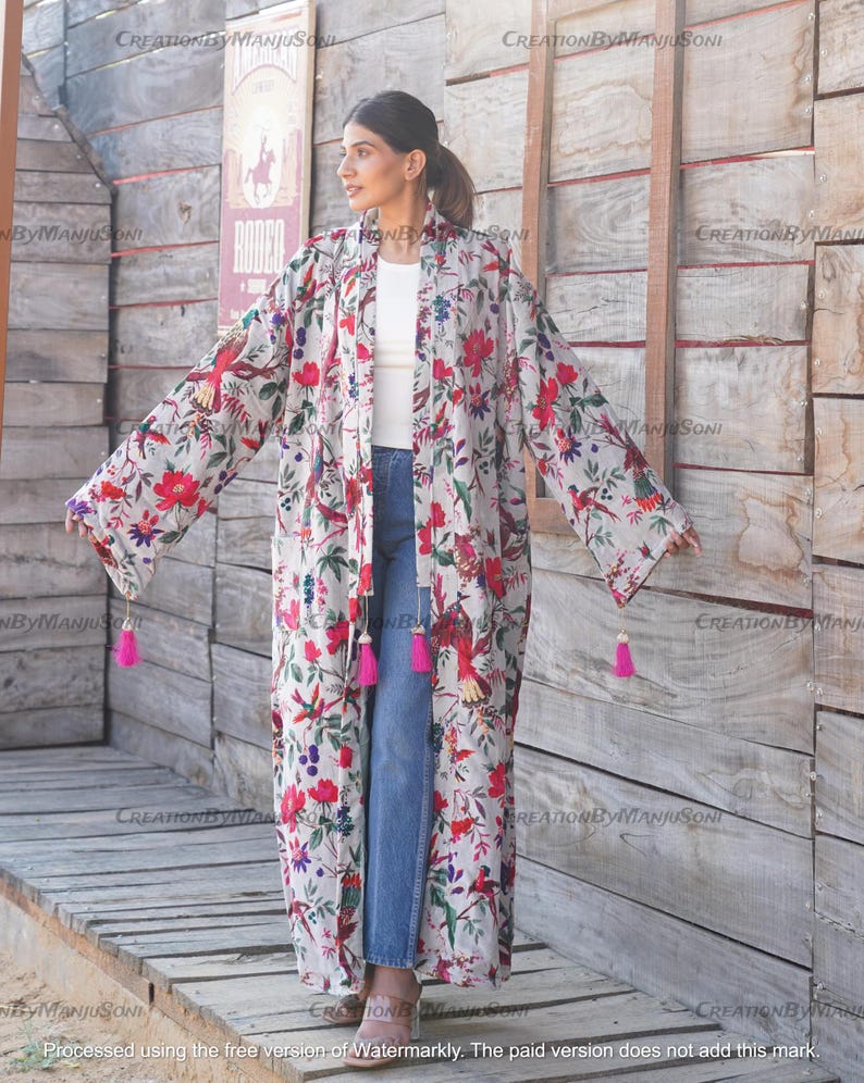May include: A long, white floral print kimono with pink tassels on the sleeves. The kimono is worn over a white t-shirt and blue jeans.