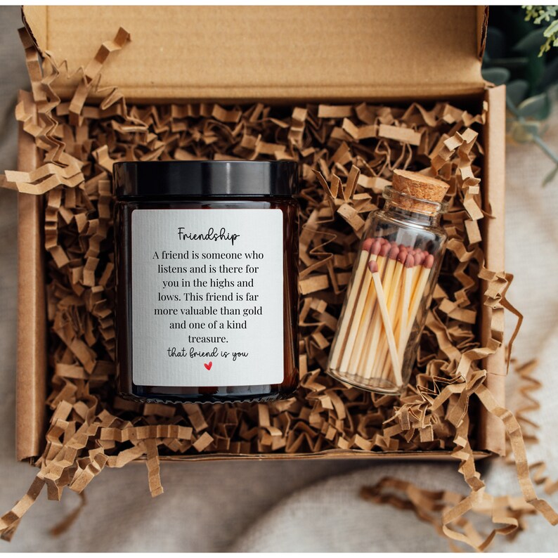 May include: A brown glass candle jar with a white label that reads "Friendship" and a quote about the value of friendship. The candle jar is in a brown cardboard box with shredded paper. A small glass jar with matches is next to the candle jar.
