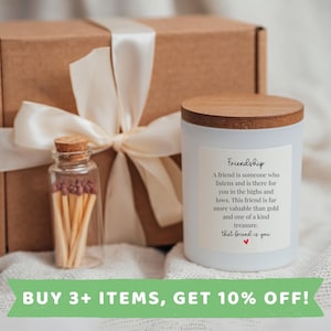 May include: White candle with a wooden lid and a label that reads "Friendship" with a quote about friendship. The candle is packaged in a brown box with a white ribbon. A small glass jar with matches is also included.