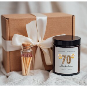 May include: A gift box with a brown paper exterior and a white satin ribbon. The box contains a black candle with a white label that says "Seventy & Fabulous" and features a gold number 70 with gold and pink balloons. The label also says "Michelle Happy Birthday to a Special Friend". A small glass jar with matches is also included.