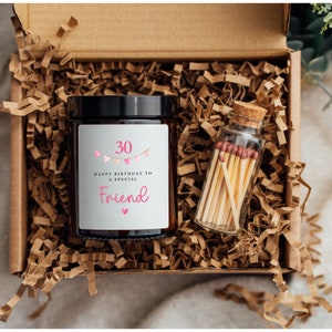 May include: A brown cardboard box filled with shredded paper. The box contains a glass jar candle with a gray label that says "30 Happy Birthday to a Special Friend" with pink bunting and a small glass jar filled with matches.