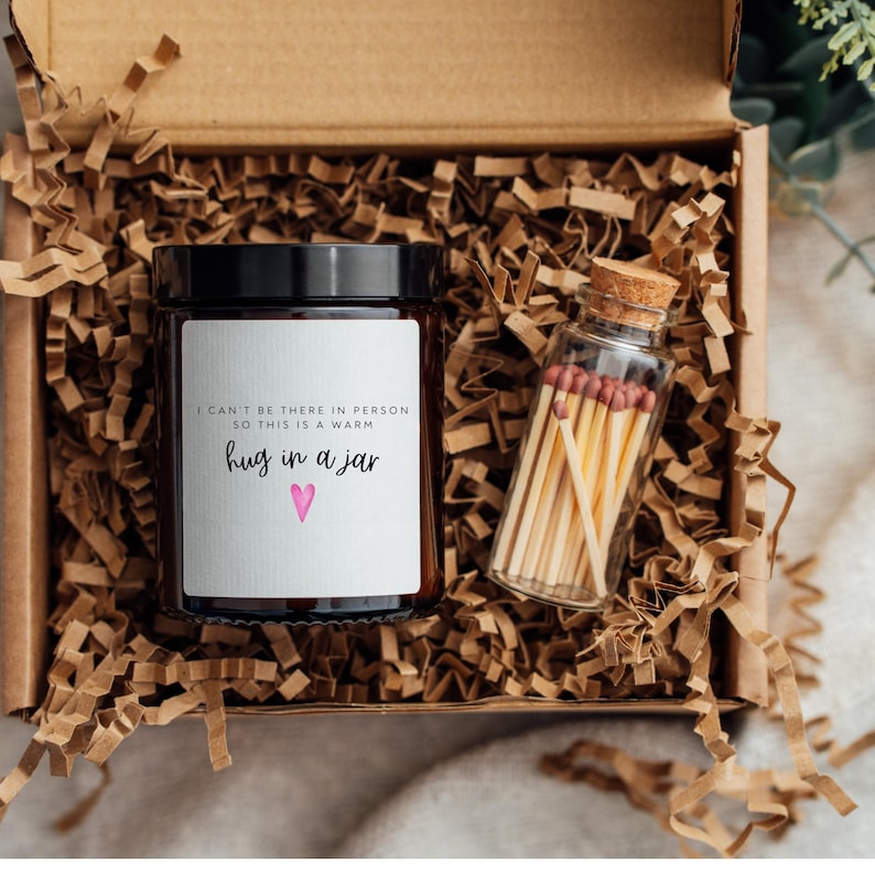 May include: A gift box filled with brown packing paper contains a glass jar candle with a white label that reads "I can't be there in person so this is a warm hug in a jar" with a pink heart.  Next to the candle is a small glass jar filled with matches.
