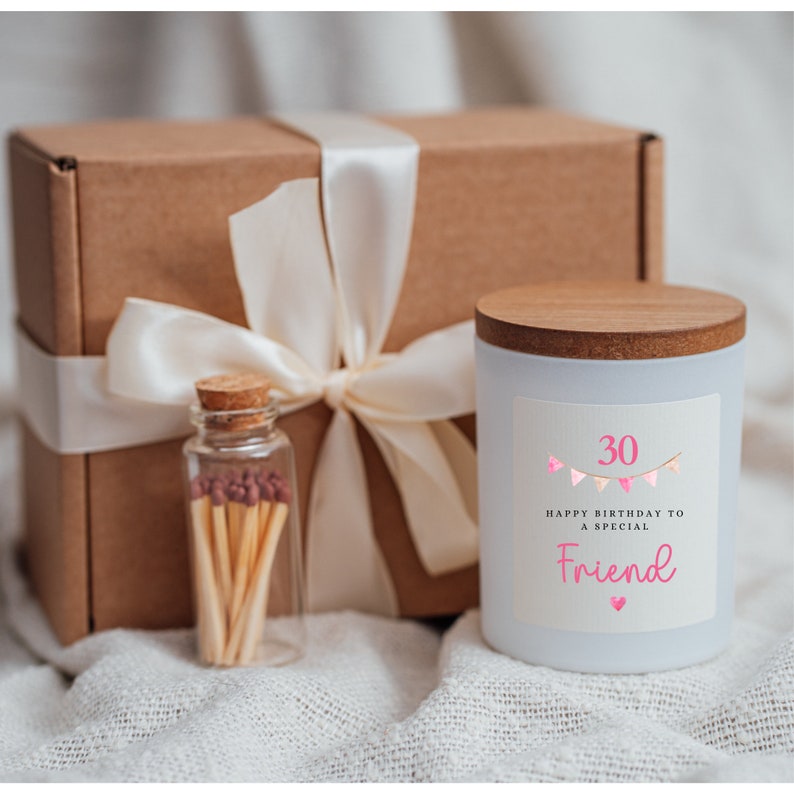 May include: A white candle with a wooden lid and a label that says "Happy Birthday to a Special Friend" with pink bunting and the number 30. The candle is in a brown box with a white ribbon and a small glass jar of matches.