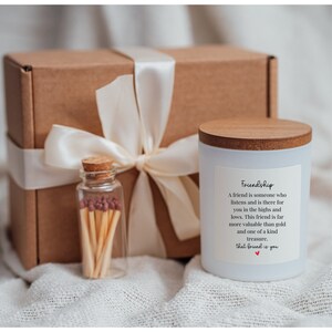 May include: A white candle with a wooden lid and a brown box tied with a white ribbon. The candle has a label that reads "Friendship" and a quote about the value of friendship. The box contains a small glass jar of matches.