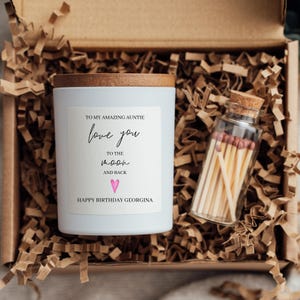 May include: A white candle in a glass jar with a wooden lid and a label reading TO MY AMAZING AUNTIE love you TO THE MOON AND BACK HAPPY BIRTHDAY GEORGINA with a pink heart, alongside a small glass bottle filled with wooden matches.
