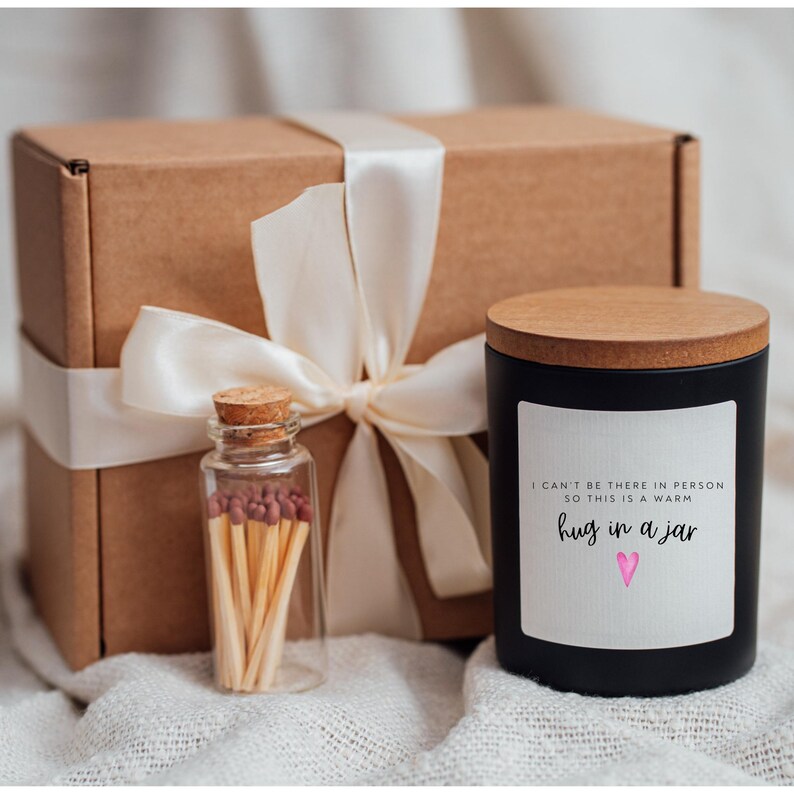May include: A black candle with a wooden lid and a white label that reads "I can't be there in person so this is a warm hug in a jar" with a pink heart. The candle is packaged in a brown box with a white ribbon and a small glass jar of matches.