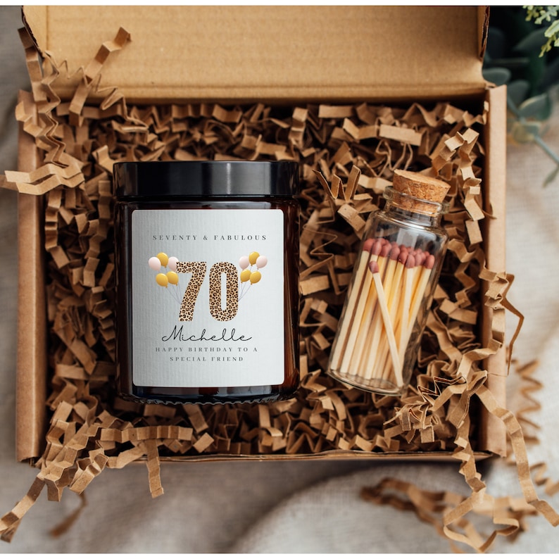 May include: A gift box filled with brown crinkle paper contains a brown glass candle with a white label that says "Seventy & Fabulous 70 Michelle Happy Birthday to a Special Friend". A small glass jar with a cork lid filled with matches is also in the box.