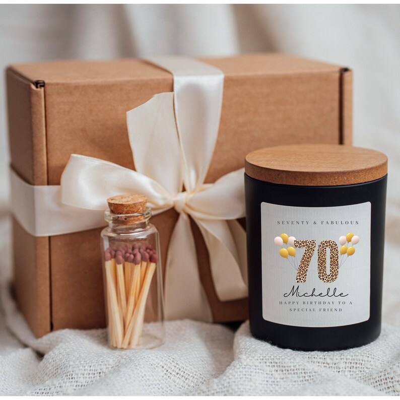 May include: A black candle with a wood lid and a label that says "Seventy & Fabulous" with a leopard print 70 and balloons. The candle is in a gift box with a white ribbon and a small glass jar of matches.