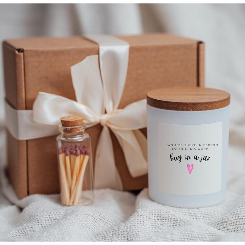 May include: A white candle in a glass jar with a wooden lid. The candle has a label that says "I can't be there in person so this is a warm hug in a jar" with a pink heart. The candle is packaged in a brown box with a white ribbon and a small glass jar of matches.