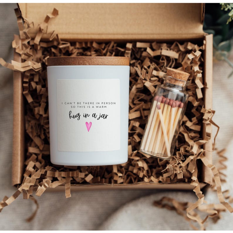 May include: A white candle jar with a wooden lid and a label that reads "I can't be there in person so this is a warm hug in a jar" with a pink heart. The candle jar is in a brown box with shredded paper and a small glass jar of matches.
