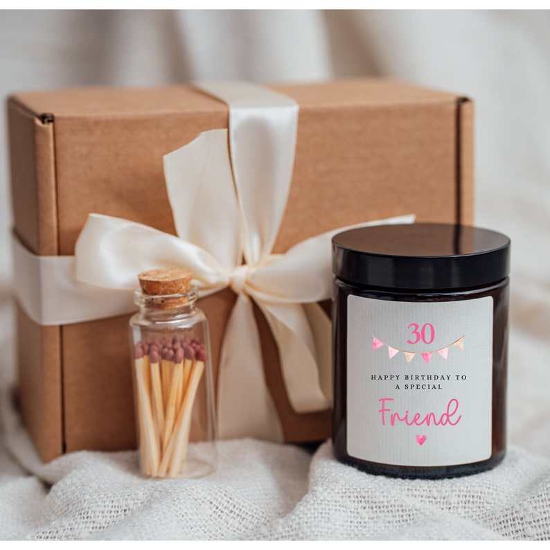 May include: A brown gift box with a white satin ribbon tied in a bow. The box contains a candle with a white label that says "Happy Birthday to a Special Friend" with pink bunting and the number 30. A small glass jar with matches is in front of the box.
