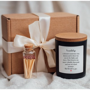 May include: A black candle with a wooden lid and a white label that reads "Friendship". The label has a quote about friendship. The candle is packaged in a brown box with a white ribbon. A small glass jar with matches is also included.