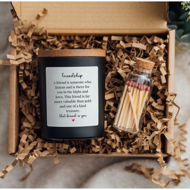 May include: A black candle with a wooden lid and a white label that reads "Friendship" with a quote about the value of friendship. The candle is in a brown cardboard box with shredded paper. A small glass jar with matches is next to the candle.