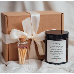 May include: A brown cardboard box with a white satin ribbon tied around it. The box contains a small glass jar of matches and a black candle with a white label that reads "Friendship. A friend is someone who listens and is there for you in the highs and lows. This friend is far more valuable than gold and one of a kind treasure. That friend is you."