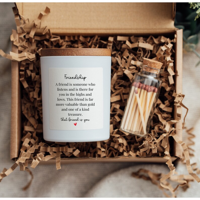 May include: A white candle with a wooden lid and a label that reads "Friendship" with a quote about the value of friendship. The candle is in a box with shredded paper and a small glass jar with matches.