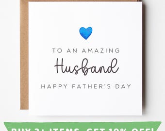 Könnte beinhalten: Eine weiße Vatertagskarte mit einem blauen Herzen und den Worten "TO AN AMAZING Husband HAPPY FATHER'S DAY". Die Karte befindet sich in einem braunen Umschlag. Der untere Teil der Karte hat ein grünes Banner mit Text.