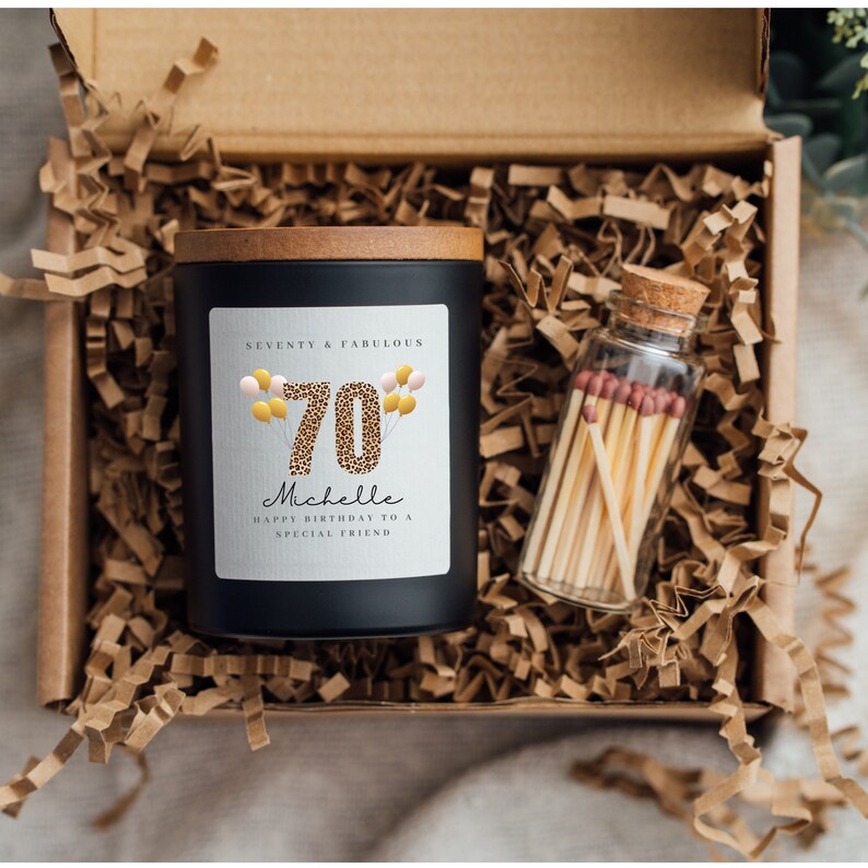 May include: A black candle with a wooden lid and a white label that says "Seventy & Fabulous" with a leopard print 70 and balloons. The candle is in a brown box with shredded paper. There is a small glass jar with matches next to the candle.