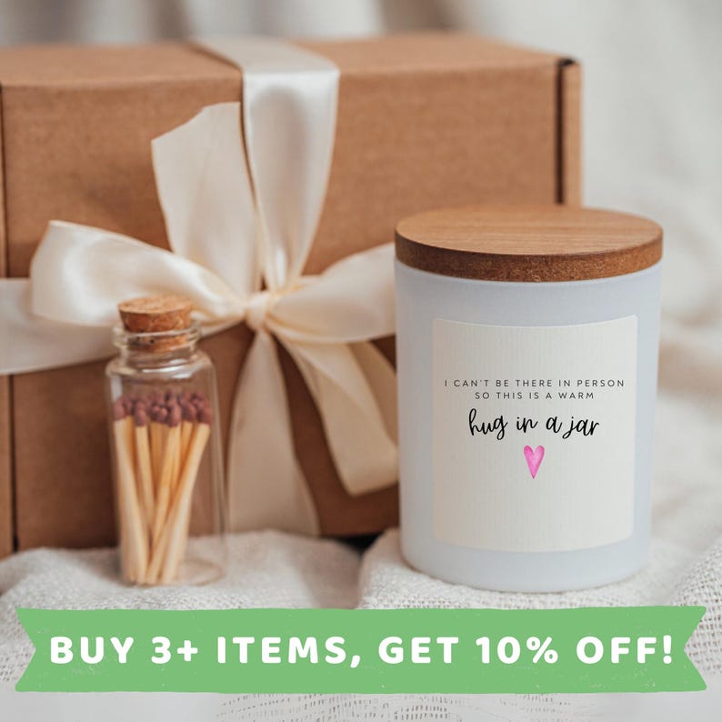 May include: A white candle with a wooden lid and a label that reads "I can't be there in person so this is a warm hug in a jar" with a pink heart. The candle is packaged in a brown box with a white ribbon and a small glass jar filled with matches.