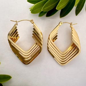 May include: A pair of gold-coloured hoop earrings. Each earring has a geometric, layered design with a square shape. The earrings are displayed on a white surface with green leaves in the background.