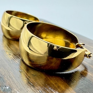 May include: Two gold-coloured hoop earrings with a polished, reflective surface. The earrings are thick and rounded, with a simple clasp closure. They are placed on a dark wooden surface, reflecting the light.