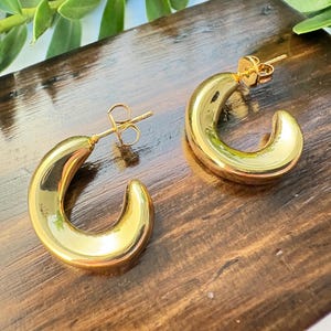 May include: A pair of gold-coloured, chunky hoop earrings. The earrings have a smooth, polished finish and a curved, crescent shape. They are displayed on a dark wooden surface, with green foliage in the background.