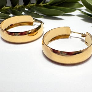 May include: A pair of gold-coloured hoop earrings. The earrings have a thick, rounded design and a shiny finish. The earrings are displayed on a white surface with green foliage in the background. The earrings are a classic jewellery item.