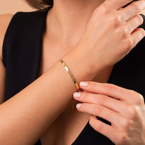 May include: A gold bracelet with a flat, textured design. The bracelet is worn on a person's wrist.