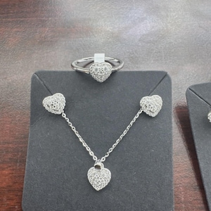 Puede incluir: Un conjunto de joyas plateadas que incluye un anillo, un collar y pendientes, todos con diseños en forma de corazón. Cada pieza está adornada con pequeñas piedras brillantes, creando una apariencia delicada y elegante. El conjunto se exhibe sobre una superficie gris oscuro.