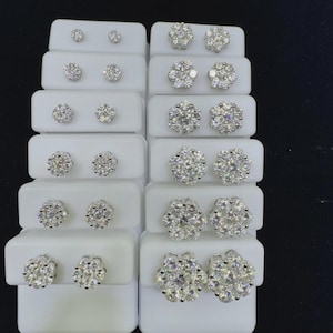 May include: A collection of diamond stud earrings displayed on white platforms. The earrings vary in size and feature a cluster design, with multiple small diamonds arranged to create a floral or starburst appearance. The background is black.