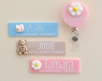 Puede incluir: Tres etiquetas de nombre de plástico rosa, azul y beige con un personaje de dibujos animados en cada una. Las etiquetas de nombre dicen "Ella Occupational Therapist", "Jodie Registered Nurse" y "Tamara Occupational Therapist". Un portagajes retráctil rosa con una margarita blanca dice "Lauren".