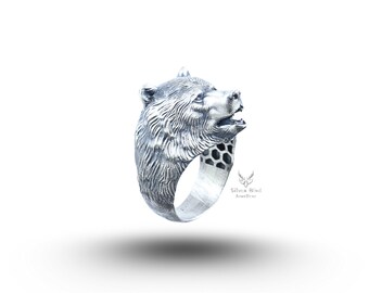 May include: A silver-colored ring featuring a detailed bear head design. The bear's face is intricately sculpted, with visible fur texture and an open mouth. The ring band has a honeycomb pattern. The ring is a piece of jewelry.