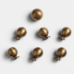 Peut inclure: Un ensemble de sept boutons de placard sphériques avec une finition en laiton antique brossé. Chaque bouton présente une tête ronde et lisse fixée sur une petite base circulaire, disposés en triangle sur un fond clair et uni.