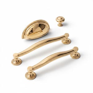 May include: A set of polished gold-tone cabinet hardware including two curved drawer pulls with beaded detailing, one semi-circular cup pull with a beaded edge, and one small round knob with a matching beaded border on a white background.