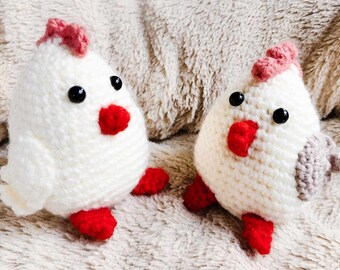 May include: Two handmade crochet chicken toys. The chickens are white with red beaks and feet, black eyes, and pink crests. The toys are sitting on a soft, textured surface.